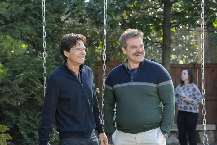 Two middle-aged men stand outdoors between the chains of a garden swing, smiling and looking off to the side. One wears glasses and a navy zip-up jacket, while the other has a thick moustache and is dressed in a green sweater with a grey stripe across the chest. They appear relaxed and mid-conversation. Behind them, slightly out of focus, a woman in a patterned jumper stands holding a drink near a wooden fence, with leafy trees and greenery filling the background.
