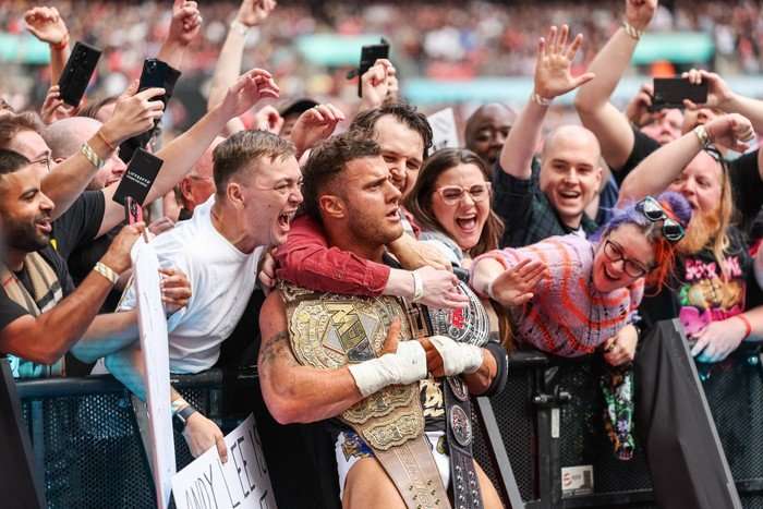 MJF draped in AEW championship belts poses in front of cheering fans at a crowded arena barrier.