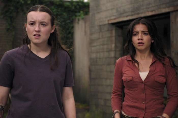 Ellie (Bella Ramsey) and Dina (Isabela Merced) in The Last of Us season 2, standing side by side outdoors in a post-apocalyptic setting. Both have tense, concerned expressions as they look offscreen. Ellie wears a dark purple t-shirt, revealing a fern tattoo on her right forearm, while Dina wears a rust-colored button-up shirt over a light tank top. The background includes weathered brick and concrete walls overgrown with greenery.
