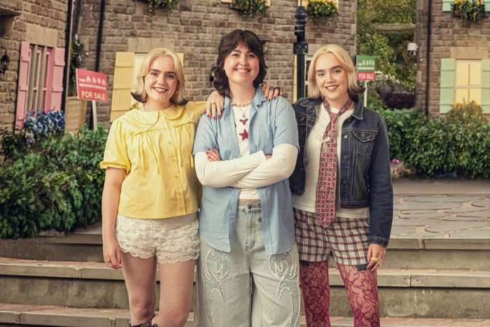 Two blonde twin sisters &ndash; one wearing a lemon top and shorts and the other wearing gingham shorts, red tights, a white shirt and tie and a denim jacket &ndash; stand on either side of a brunette woman &ndash; who is wearing an open shirt over a long-sleeve white shirt and trousers and has her arms crossed, all three are smiling for the camera.