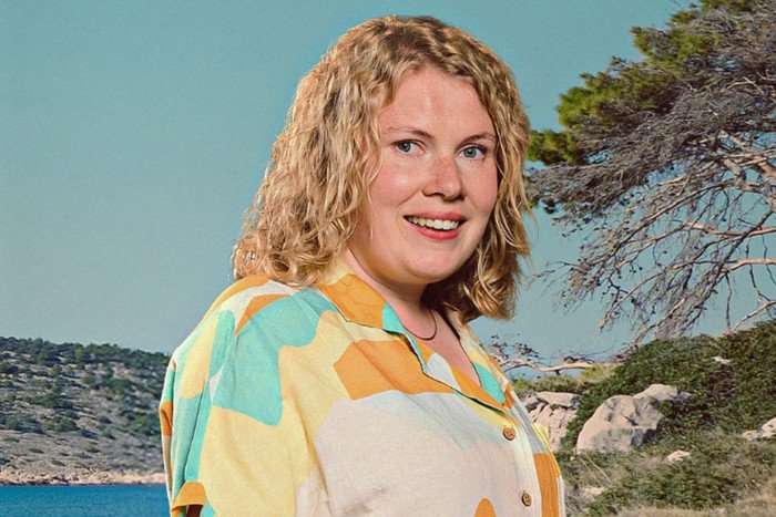 A woman with shoulder length curly blonde hair smiles for the camera. She's wearing a multicoloured button-up shirt