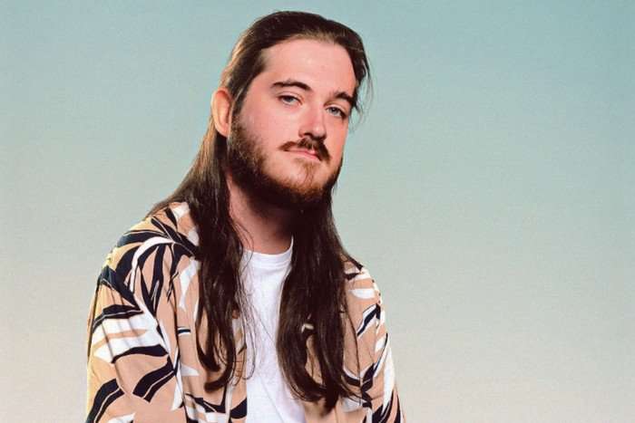 A man with long brown hair and a beard poses for the camera. He's wearing a white t-shirt under a cream and black overshirt