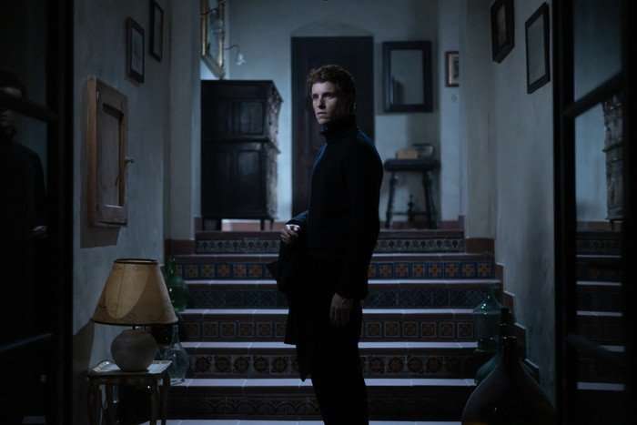 The Jackal stands in a room of his house, with all the lights turned off, looking ahead of him severely