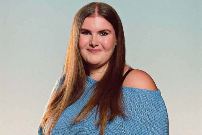 A woman with long straight brown hair smiles as she poses for the camera. she's wearing a blue off-shoulder top
