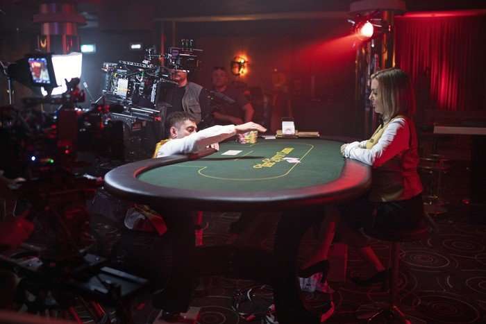 Michael Socha and Sheridan Smith being filmed on the set of The Cage, in a darkened casino room, both leaning on a baize topped card table.