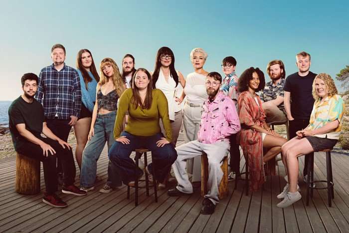 14 people stand collectively against a clear blue sky on what appears to be a wooden boardwalk