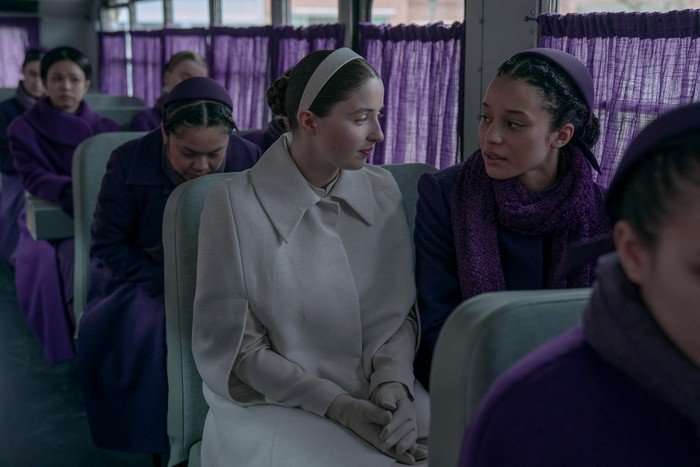 A woman in white and a woman in purple sit on a bus talking. The windows of the bus are draped in purple curtains