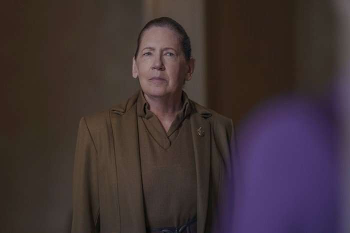Ann Dowd as Aunt Lydia in the Testaments, wearing a brown outfit and staring suspiciously at someone stood in front of her