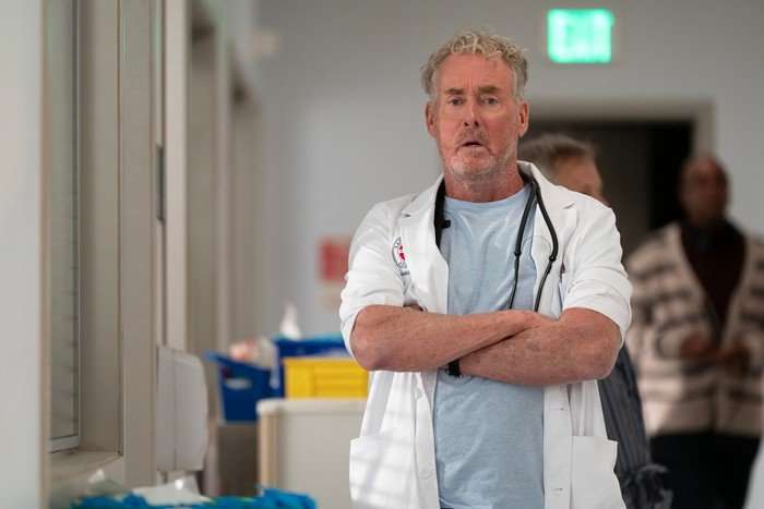 John C McGinley stars in Scrubs season 10; Dr Cox is standing with his arms crossed, looking down a hospital corridor with an unimpressed facial expression
