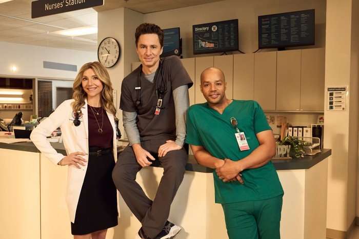 Scrubs stars Sarah Chalke as Elliot Reid, Zach Braff as John 'JD' Dorian, and Donald Faison as Christopher Turk &ndash; they are stood together at the reception desk of a Sacred Heart hospital ward.