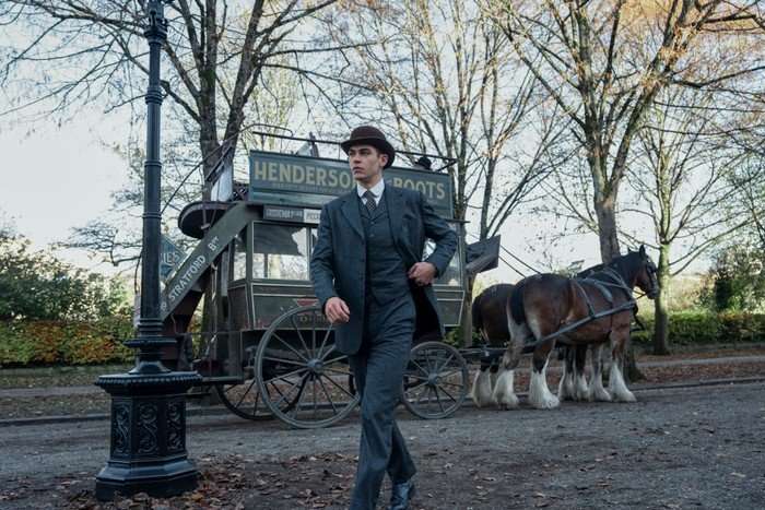 Hero Fiennes Tiffin as Sherlock Holmes in Young Sherlock, wearing a suit and hat and walking outside by a horse and carriage.