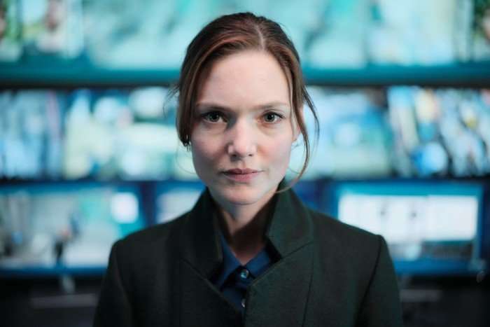 Holliday Grainger as Rachel Carey in The Capture, stood looking at the camera with a set of screens behind her.