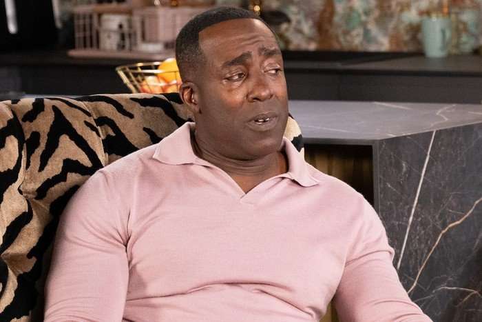 Ronnie, in a pink shirt, sits beside Carl, in a blue shirt, sipping whiskey in Debbie's flat in Coronation Street