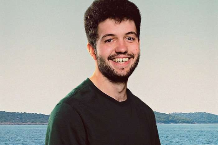 A man with puffy dark brown hair, a short beard and moustache smiles for the camera, he's wearing a black t-shirt