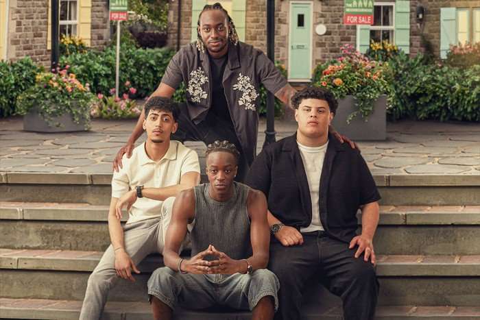 Four boys, aged between 20 and 21, pose together in a town square. All four have straight faces, two in black jeans and two in light blue jeans.