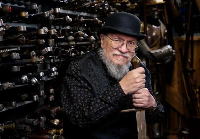 418734_Game of Thrones_ The Mad King_2026 George RR Martin wearing a black shirt and black bowler hat, leaning on a large sword and looking at the camera.