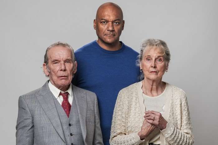 George Knight, in a blue jumper, stands behind Eddie and Gloria Knight in key art for EastEnders. Eddie is wearing a grey suit and red tie, and Gloria has a beige cardigan on.