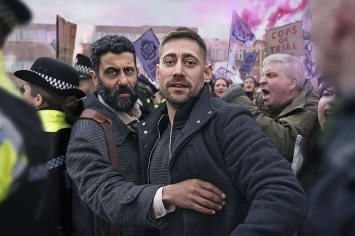Two men standing in a protest, one stretching his arm out in front of the other