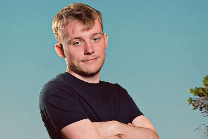 A man with strawberry blond hair has his arms folded while lightly smiling for the camera. he's wearing a black t-shirt