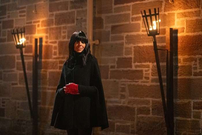 Claudia Winkleman in The Traitors against a brick wall with two fire-lit lamps wearing red ringerless gloves and a black cape.