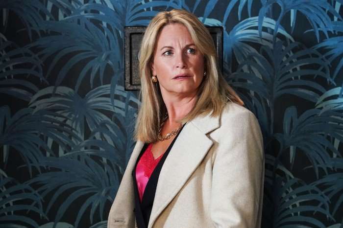 A shocked Sam Mitchell stands in a white coat against a blue wallpapered wall in EastEnders