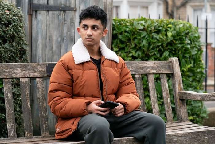 Nugget looks stunned, sat in a padded orange coat on Arthur's bench in a scene from EastEnders