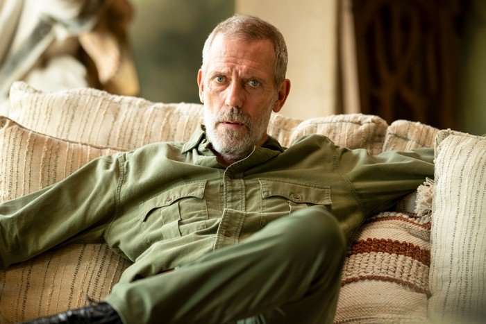 Richard Roper sits on a sofa in his home, wearing a green shirt and trousers, staring ahead with concern