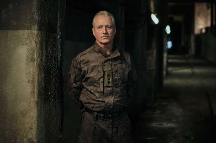 Linus Roache as Colonel Figgis in The Capture, wearing a military uniform and stood in a dark corridor.