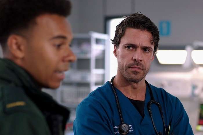 A man dressed in scrubs and a stethoscope around his neck looks seriously at another man dressed as a paramedic and out of focus.