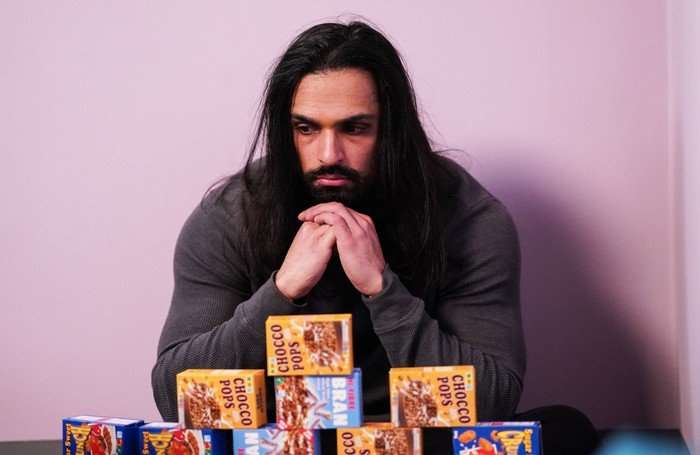 Ravi sits in front of some stacked cereal boxes in a scene from EastEnders