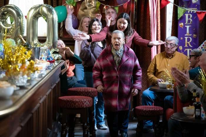 Mo walks into The Vic surrounded by friends and family, with a shocked expression on her face and some big balloons with the number '90' on them in a scene from EastEnders