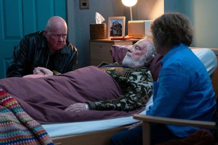 Phil and Julie sit beside Nigel, who is asleep in bed, in a scene from EastEnders