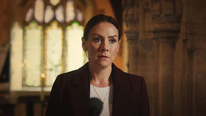 Stevie stands in a church in a scene from Casualty, wearing a white blouse and black jacket.