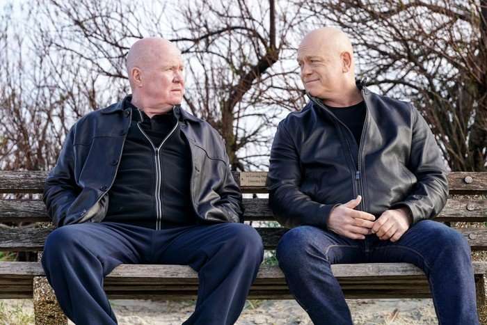 Phil and Grant sit on a bench looking towards one another in a scene from EastEnders