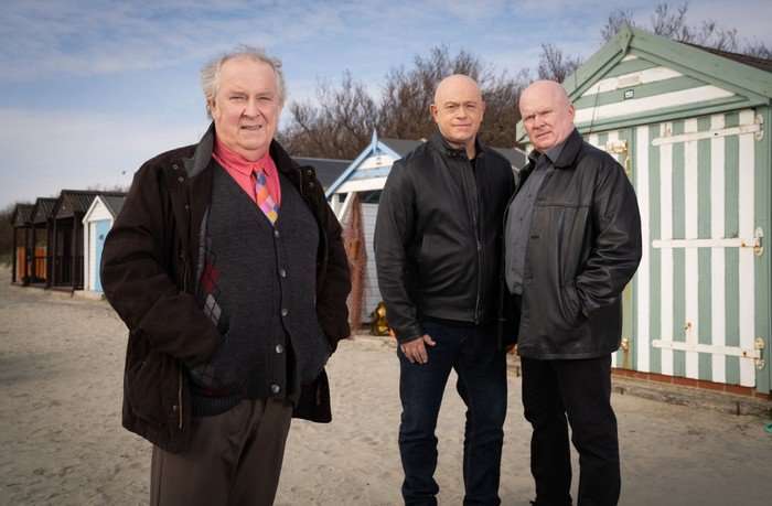 Nigel, Phil and Grant stare into the camera on a sandy beach in front of some beach houses in a scene from EastEnders