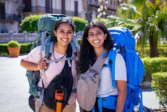 Puja and Roshni stood next to each other smiling ahead with large rucksacks on their backs.