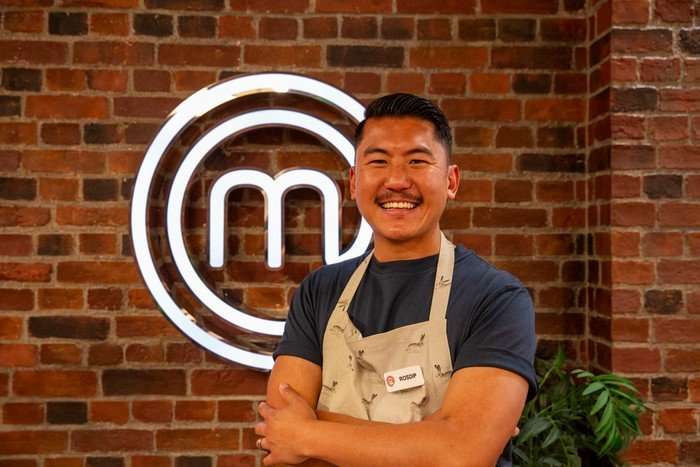 Mat is in a navy blue t-shirt and beige apron, and poses with his arms crossed in front of a brick wall bearing the MasterChef logo