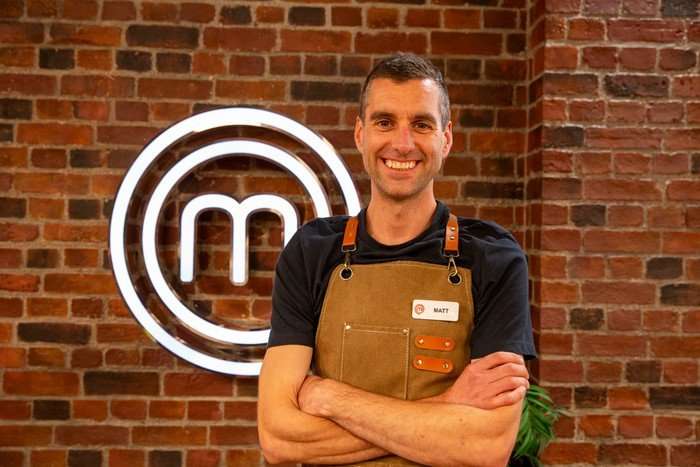 Mat is in a black t-shirt and brown apron, and poses with his arms crossed in front of a brick wall bearing the MasterChef logo