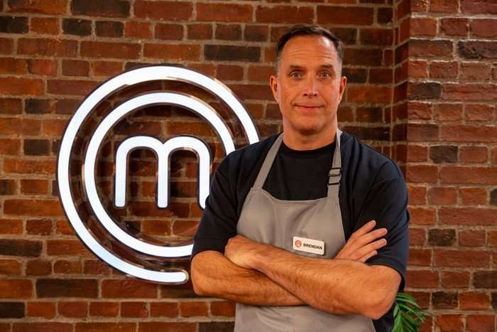 A man in a blue t-shirt and grey apron poses with his arms crossed in front of a brick wall bearing the MasterChef logo