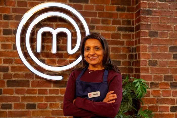 Sabina is in a maroon long-sleeved shirt and navy blue apron, and poses with her arms crossed in front of a brick wall bearing the MasterChef logo