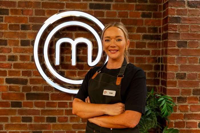 A woman poses with her arms crossed in front of a brick wall bearing the MasterChef logo. She is wearing a navy-blue t-shirt and a black apron, and has her blonde hair swept up in a bun.