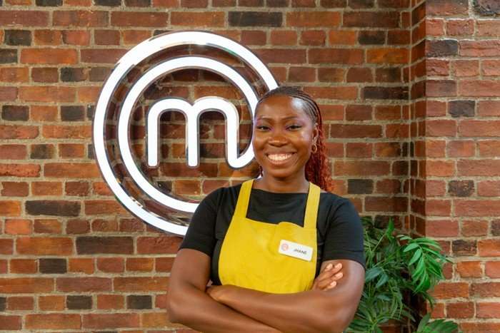 A woman poses with her arms crossed in front of a brick wall bearing the MasterChef logo. She is wearing a black shirt and a yellow apron, and has a big smile on her face, with her brown and red hair pulled back with head braids