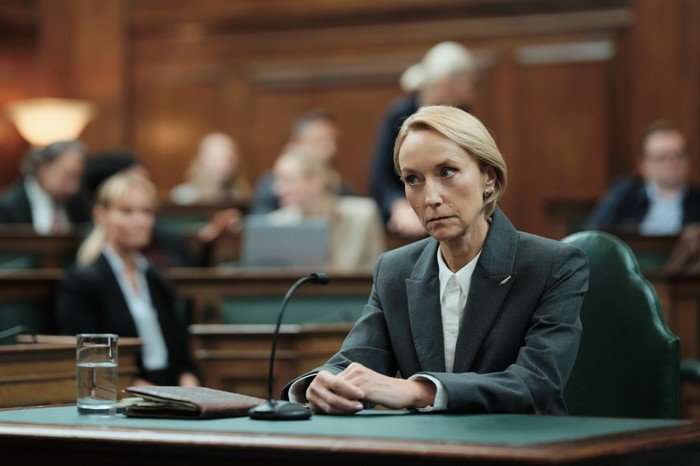 Lia Williams as Gemma Garland in The Capture, sat in parliament behind a green desk, with a microphone in front of her and people sat behind her.