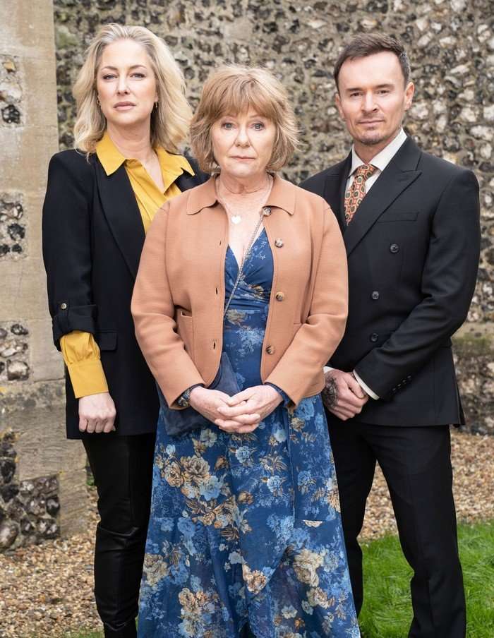 Clare wearing a black blazer and yellow shirt, stood beside Julie in a brown jacket and blue dress and Josh in a black suit at Nigel's funeral in EastEnders