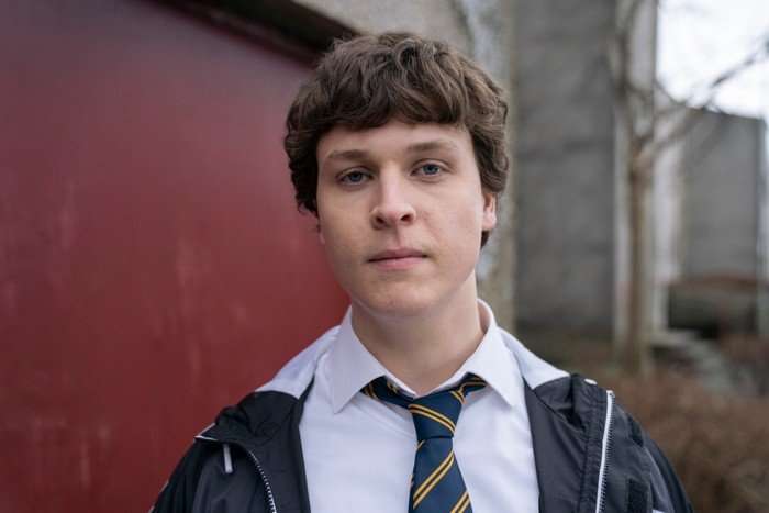 Calum Manchip as Drew in Half Man, wearing a school uniform and black jacket, stood outside by a garage.