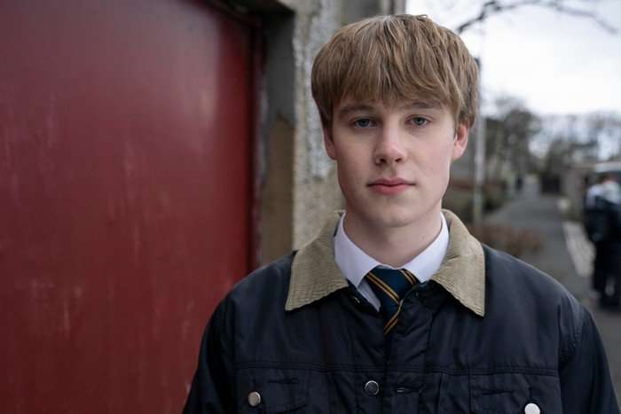 Piers Ewart as Young Gus in Half Man, wearing a school uniform and coat, stood outside by a garage.