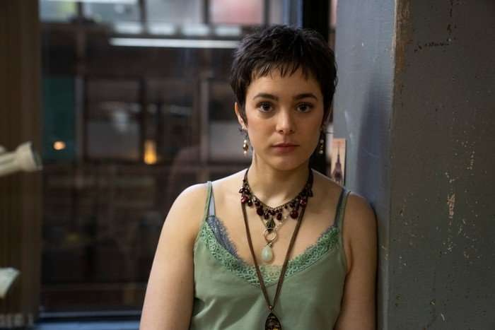 Philippine Velge as Celeste in Half Man, wearing a green vest and a necklace, stood against a wall.