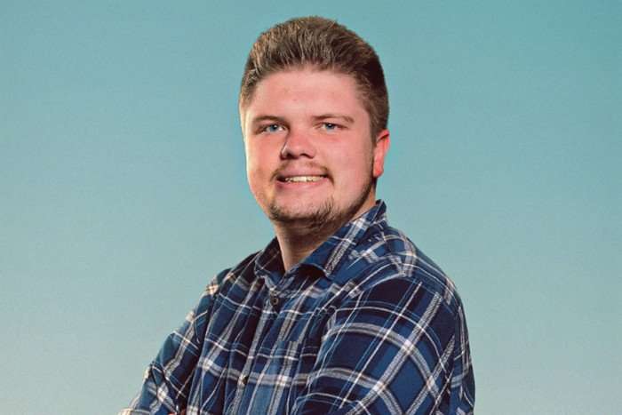 A man with dark blond hair smiles for the camera, he's wearing a blue gingham shirt