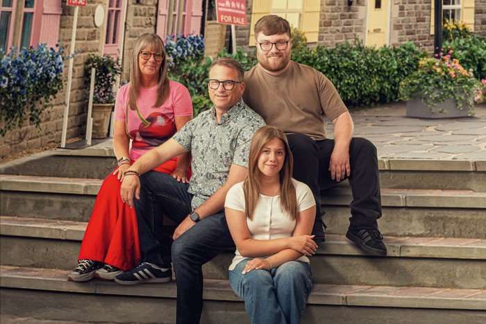 A family of four are sitting on steps in a town square. Wendy, on the left, is wearing a pink shirt and red trousers, with husband Paul in a floral shirt next to him, leaning on his knee. Behind him is son Harrison, who is in a brown shirt and black trousers, and in front of him is daughter Grace, who is in a white shirt and jeans with her arms crossed over her stomach as she sits