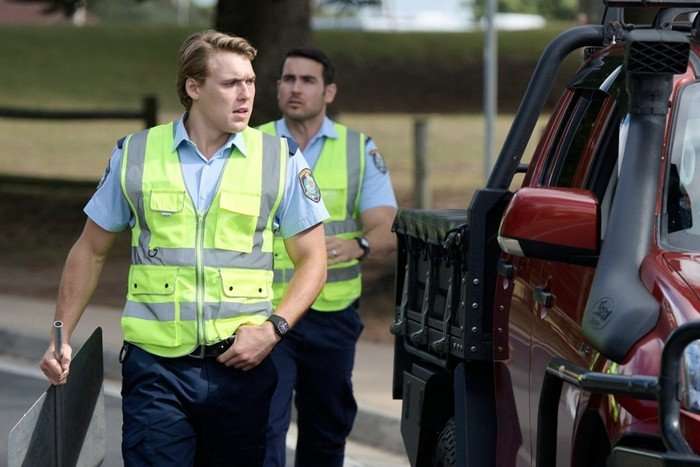 Ricchie and Cash, wearing high vis jackets, walk along a road beside a red car in Home and Away